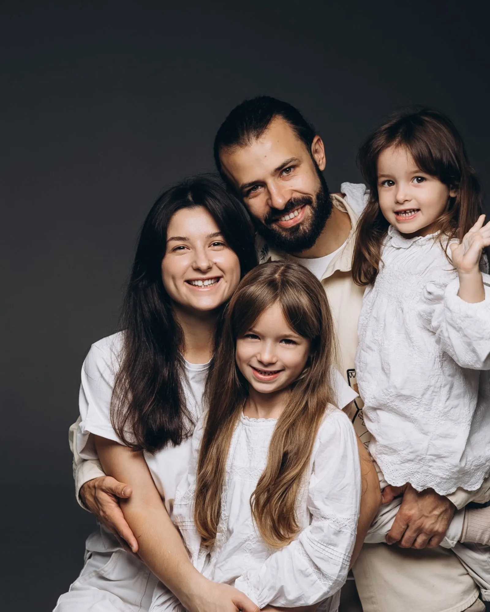 A family of four in white, smiling together against a dark studio backdrop
